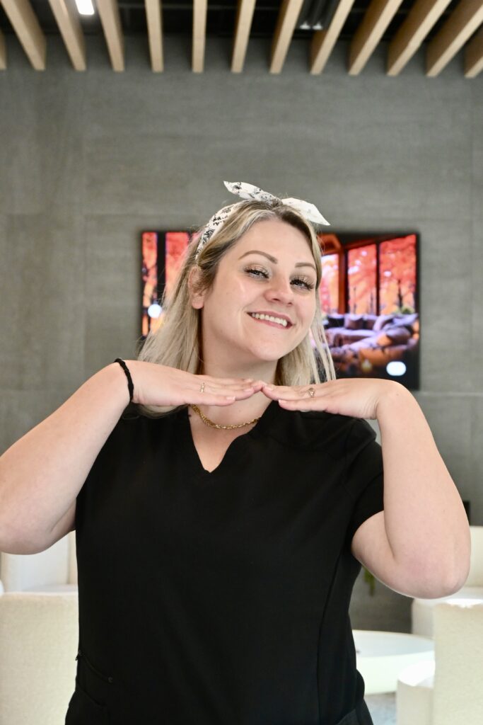 Jamie Masefield, dental assistant at Premier Prosthodontics, smiling playfully in black scrubs inside the practice lobby.